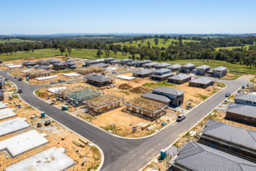 Aerial view of a new suburban subdivision under construction, with slabs and framed homes at various stages and rolling hills behind